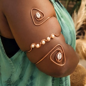 May include: A copper wire arm cuff featuring two triangular shapes, each centered with a white cowrie shell, connected by a central wavy wire section adorned with four small, round, cream-colored pearls arranged in a linear pattern.