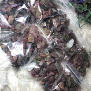 May include: Multiple small bags filled with pink, green, and black rough gemstones. The bags are arranged on a white fluffy surface.
