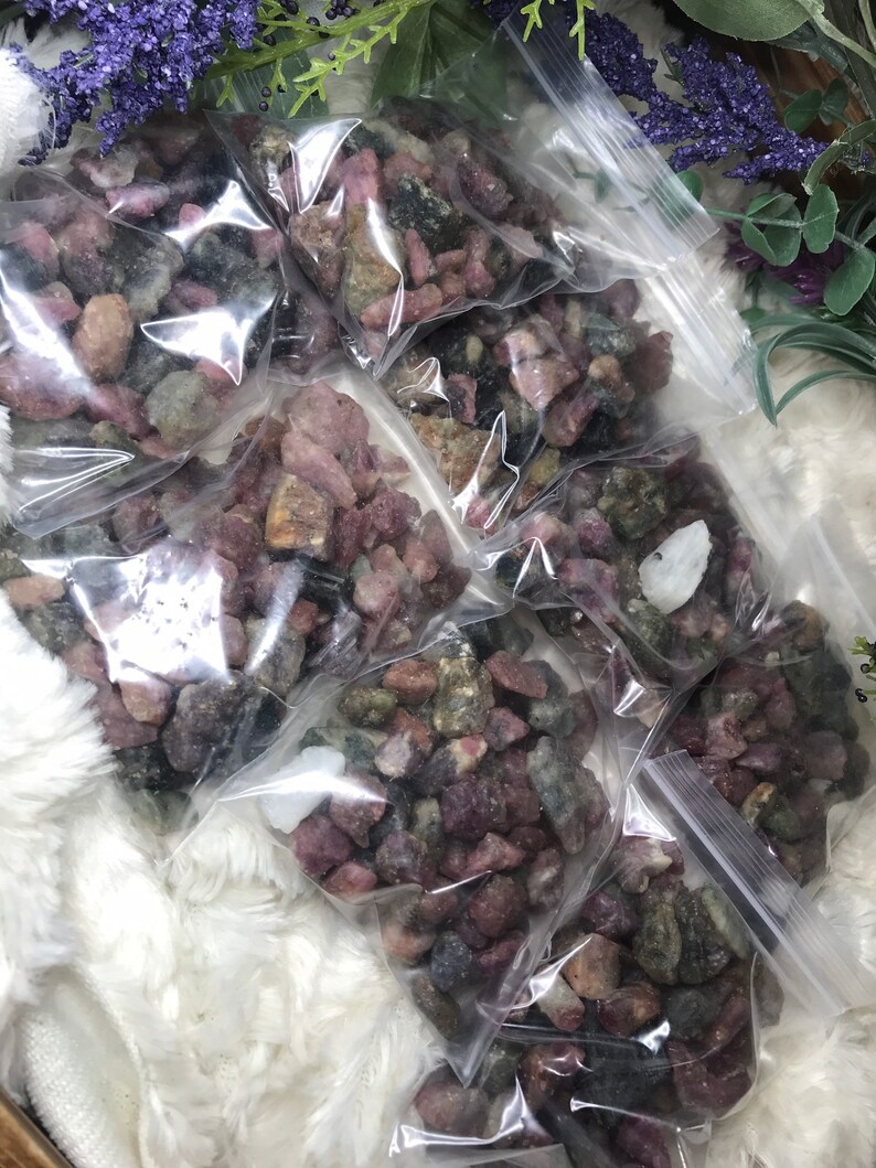 May include: Multiple small bags filled with pink, green, and black rough gemstones. The bags are arranged on a white fluffy surface.