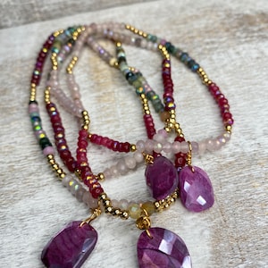 May include: A layered necklace with multiple strands of beads in shades of pink, red, and green. The necklace features three large, faceted purple gemstone pendants.