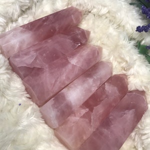 May include: Five pink rose quartz crystal points, also known as wands, are arranged in a pile on a white fluffy surface. The points are smooth and polished, with a natural, cloudy appearance.