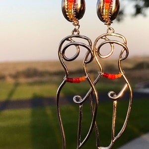 May include: A pair of silver earrings featuring a stylized human figure with a beaded waistline. The earrings are suspended from a seashell charm with a beaded design in shades of red, orange, and yellow.