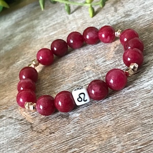 Puede incluir: Pulsera de piedras preciosas rojas con detalles dorados y un dije blanco con el símbolo astrológico de Leo.