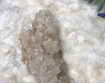 Op de afbeelding: Een cluster van heldere, witte en lichtbruine kristallen met een ruwe, natuurlijke textuur. De kristallen zijn in een cluster gerangschikt, die lijkt op een klein gebergte. De kristallen bevinden zich op een wit, pluche oppervlak.