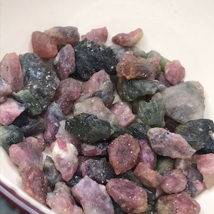 May include: A bowl filled with a variety of pink, green, and black gemstones. The gemstones are rough and have a natural, unpolished look.