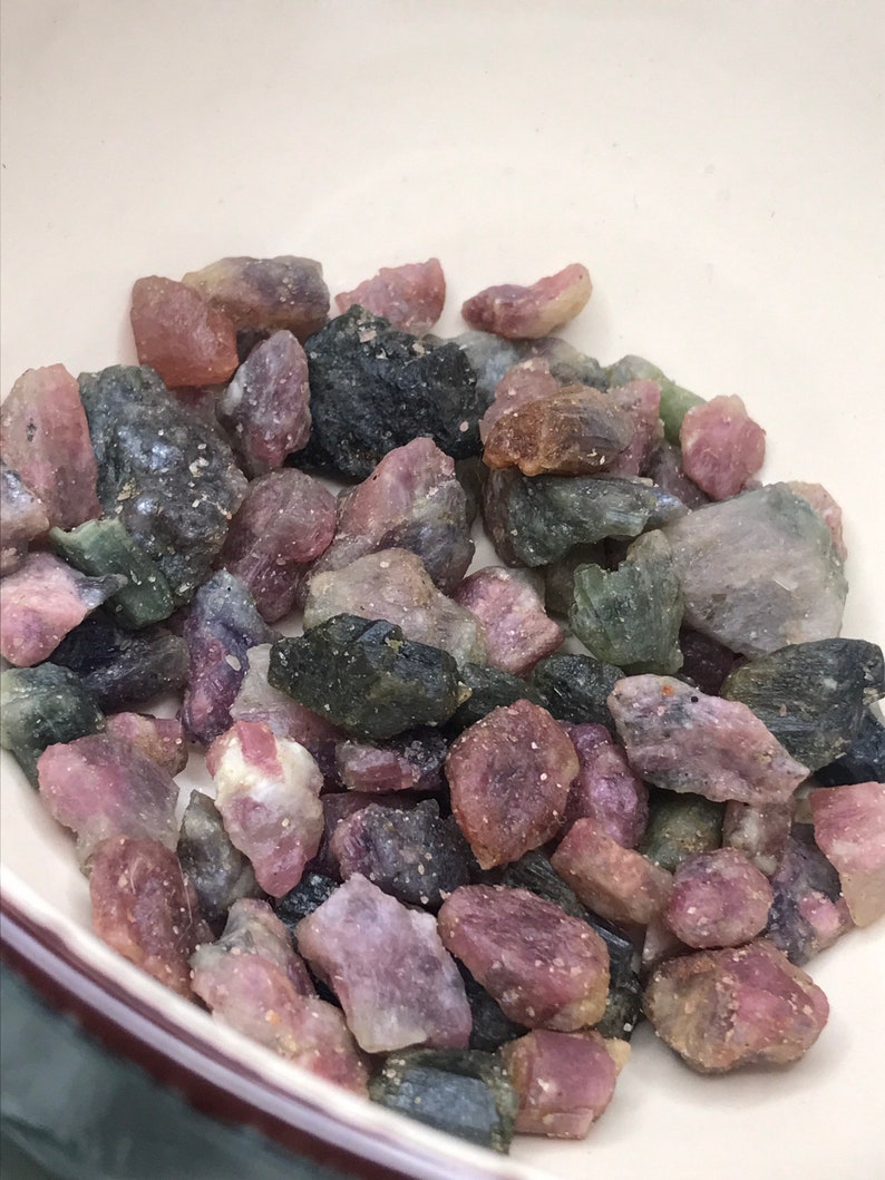 May include: A bowl filled with a variety of pink, green, and black gemstones. The gemstones are rough and have a natural, unpolished look.