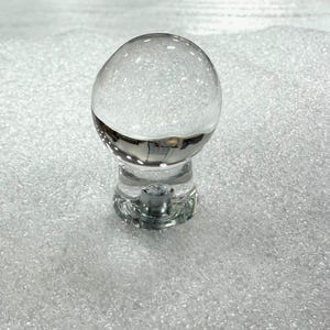 May include: A clear glass cabinet knob featuring a spherical top and a cylindrical base. The knob has a smooth, transparent surface with a metallic attachment point visible at the center of the base, designed for mounting onto furniture or cabinetry.