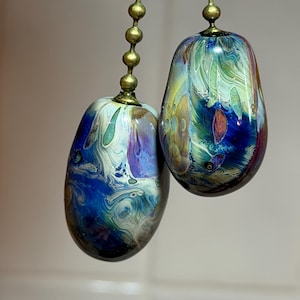 May include: Two oval-shaped glass ceiling fan pulls featuring a swirling marbled pattern of deep blue, teal, purple, and white colors. Each glass pendant is attached to a metallic brass-toned ball chain connector at the top, hanging vertically.
