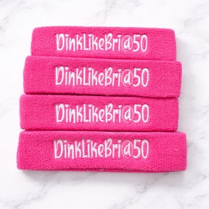 Puede incluir: Cuatro cintas para la cabeza de color rosa brillante con texto bordado en blanco "DinkLikeBri@50". Las cintas est&aacute;n apiladas sobre una superficie de m&aacute;rmol blanco y gris. Ideales para deportes o fitness.
