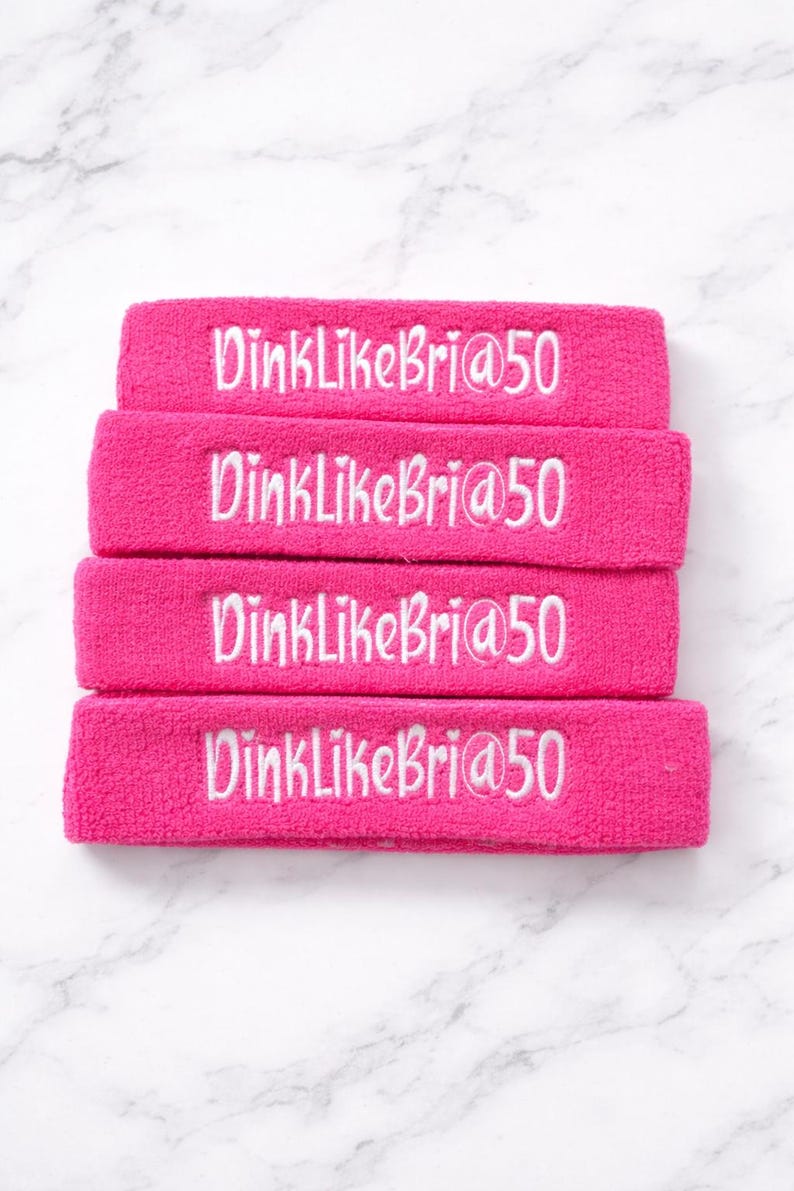 Puede incluir: Cuatro cintas para la cabeza de color rosa brillante con texto bordado en blanco "DinkLikeBri@50". Las cintas est&aacute;n apiladas sobre una superficie de m&aacute;rmol blanco y gris. Ideales para deportes o fitness.