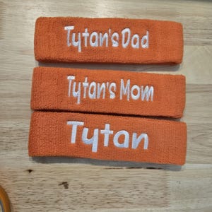 Puede incluir: Tres cintas para la cabeza de rizo naranja con texto bordado en blanco. La cinta superior dice "Tytan's Dad", la del medio "Tytan's Mom" y la inferior "Tytan".