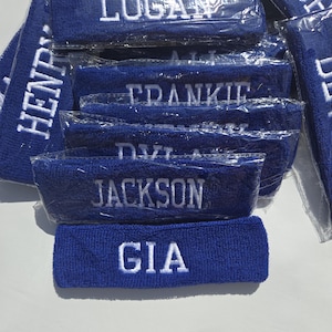 Puede incluir: Un conjunto de cintas para la cabeza de rizo azul, cada una bordada con un nombre en hilo blanco. Se ven nombres como "LOGAN", "JACKSON" y "GIA". Las cintas est&aacute;n envueltas individualmente en pl&aacute;stico transparente.
