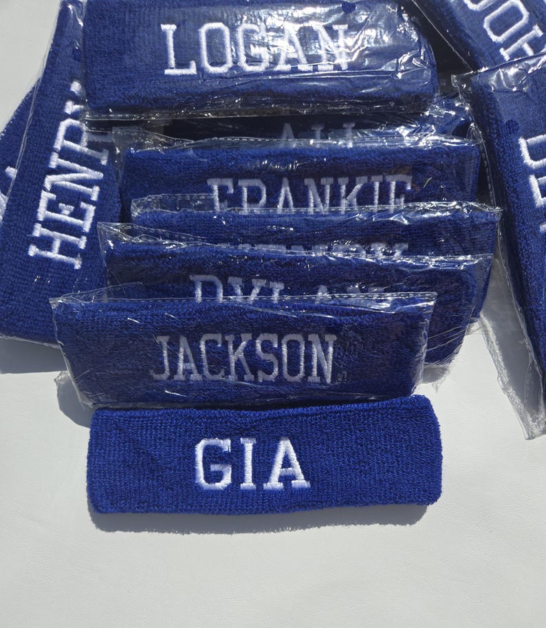 Puede incluir: Un conjunto de cintas para la cabeza de rizo azul, cada una bordada con un nombre en hilo blanco. Se ven nombres como "LOGAN", "JACKSON" y "GIA". Las cintas est&aacute;n envueltas individualmente en pl&aacute;stico transparente.