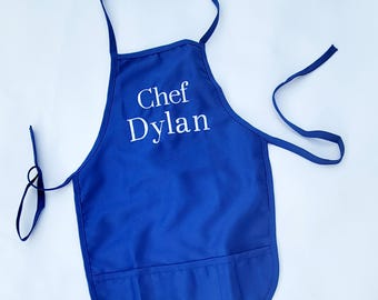 Könnte beinhalten: Blaue Kochschürze mit einer weißen Tasche und dem gestickten Text "Chef Dylan" auf der Vorderseite.