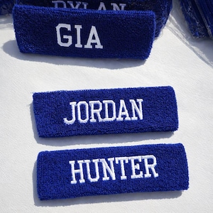 Puede incluir: Tres cintas para la cabeza de rizo azul con nombres bordados en blanco: GIA, JORDAN y HUNTER. Las cintas son rectangulares y parecen nuevas. El fondo es una superficie blanca.