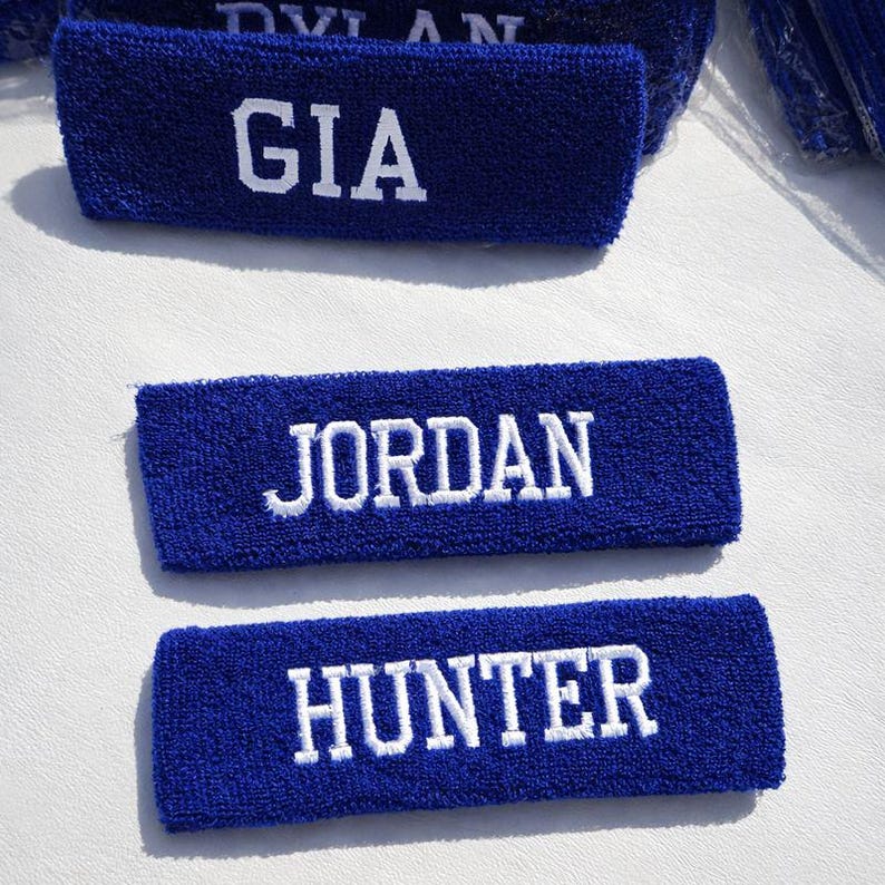 Puede incluir: Tres cintas para la cabeza de rizo azul con nombres bordados en blanco: GIA, JORDAN y HUNTER. Las cintas son rectangulares y parecen nuevas. El fondo es una superficie blanca.