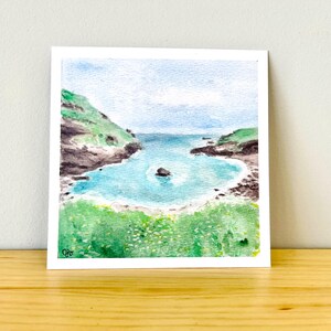 May include: Watercolor painting of a coastal scene. The artwork depicts a turquoise bay surrounded by green hills and rocky cliffs. The sky is a light blue, and the painting is on a white square card.