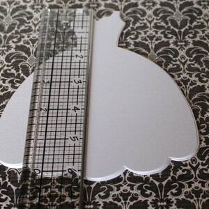 Pu&ograve; includere: Una forma di abito bianco in carta ritagliata, che misura circa 15 cm di lunghezza, su uno sfondo a motivi bianco e nero.