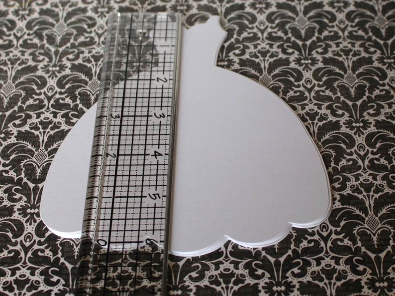 Pu&ograve; includere: Una forma di abito bianco in carta ritagliata, che misura circa 15 cm di lunghezza, su uno sfondo a motivi bianco e nero.