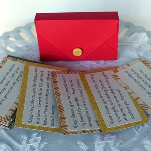 Puede incluir: Una caja de sobre roja con cierre de bot&oacute;n, que contiene una pila de tarjetas peque&ntilde;as con rayas amarillas y naranjas. Las tarjetas tienen mensajes inspiradores sobre Dios y Jes&uacute;s.