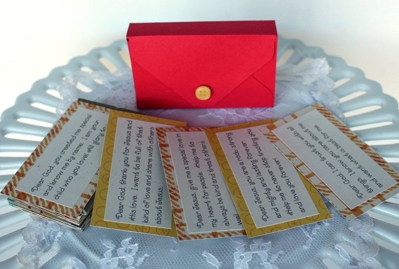 Puede incluir: Una caja de sobre roja con cierre de bot&oacute;n, que contiene una pila de tarjetas peque&ntilde;as con rayas amarillas y naranjas. Las tarjetas tienen mensajes inspiradores sobre Dios y Jes&uacute;s.