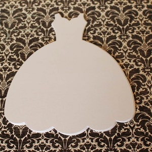 Pu&ograve; includere: Taglio di carta bianco a forma di abito da sposa. Il taglio &egrave; su uno sfondo marrone a motivi.