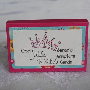Puede incluir: Una caja rosa con un dise&ntilde;o floral azul y blanco. La caja tiene una etiqueta blanca con una corona rosa y el texto "God's little PRINCESS Cards" y "Sarah's Scripture".