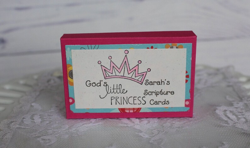 Puede incluir: Una caja rosa con un dise&ntilde;o floral azul y blanco. La caja tiene una etiqueta blanca con una corona rosa y el texto "God's little PRINCESS Cards" y "Sarah's Scripture".