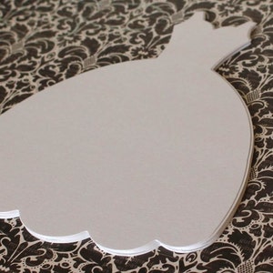 Pu&ograve; includere: Sagoma di abito bianco in carta, a forma di abito da sposa, su uno sfondo a motivi bianco e nero.