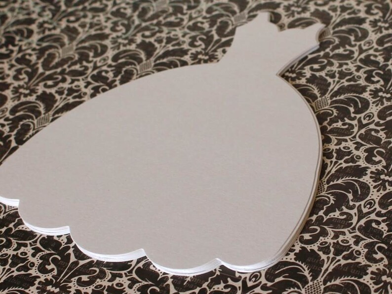 Pu&ograve; includere: Sagoma di abito bianco in carta, a forma di abito da sposa, su uno sfondo a motivi bianco e nero.