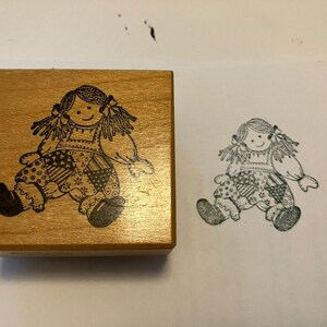 May include: A square wooden rubber stamp featuring a black line art illustration of a doll with yarn-style hair, a dress with a patchwork pattern, and boots. The stamp is shown next to a black ink impression of the same doll design on paper.