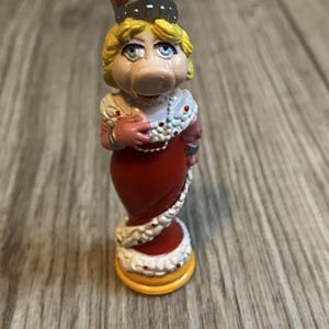 May include: A figurine of Miss Piggy from The Muppets, wearing a red dress with white trim and a silver crown. The figurine has blonde hair, blue eyes, and a pink snout. The base is yellow.