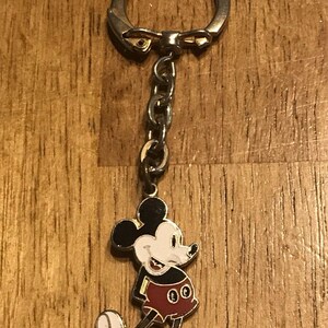 May include: A Mickey Mouse keychain featuring a smiling Mickey in his classic red shorts, black jacket, and white gloves and shoes. The keychain has a silver-colored ring and chain.