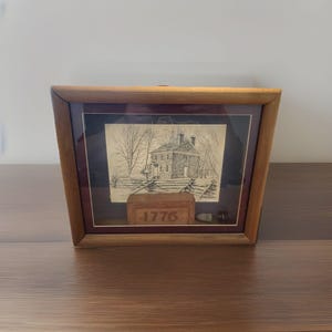 May include: Wooden framed artwork with a black and white sketch of a colonial house and fence. The frame has a dark red mount. A small brick with the year "1776" is in front of the artwork. The frame is made of wood.
