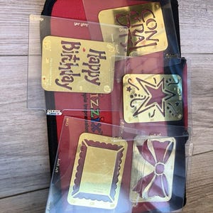May include: A selection of gold-coloured greeting card inserts with different designs and messages, including "Happy Birthday" and "Congrats." The inserts are in clear plastic sleeves, set against a red and black background.