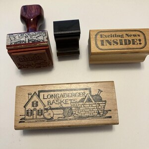 Può includere: Quattro timbri in gomma con manici o blocchi in legno. Un timbro presenta una grafica con casa e cestino con il testo LONGABERGER BASKETS. Un altro timbro riporta la scritta Exciting News INSIDE!. Un terzo timbro reca il testo I LOVE Baskets. Un righello è visibile.