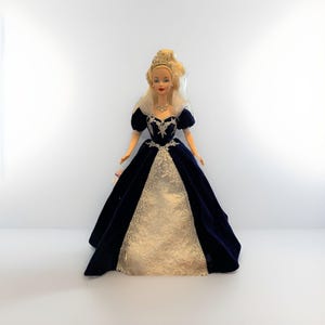 May include: A doll wearing a dark blue velvet gown with a cream-colored lace panel. The doll has blonde hair, a tiara, and a delicate necklace. The gown features short sleeves and a high collar.