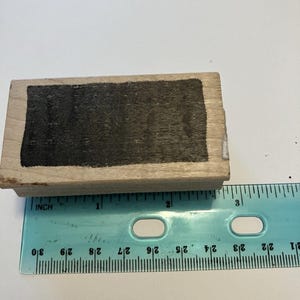 May include: A rectangular wooden rubber stamp with a black ink pad surface, positioned above a light blue plastic ruler. The stamp measures approximately 2.75 inches in length, as indicated by the markings on the ruler placed directly below it.