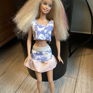May include: A doll with long blonde hair, blue eyes, and a smiling expression. The doll wears a two-piece outfit: a blue and white patterned crop top and a pink skirt with a white waistband. The doll is standing on a wooden surface.