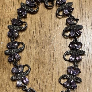 Puede incluir: Una pulsera plateada con un diseño floral y detalles de piedras preciosas moradas. La pulsera tiene un cierre decorativo y está compuesta por múltiples eslabones en forma de flor. Las piedras son ovaladas.