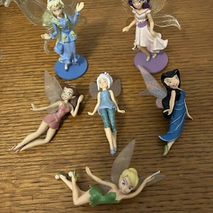May include: A collection of six fairy figurines with translucent wings. The fairies are in various poses and wear dresses in shades of blue, pink, green, and white. Each figurine is on a small, colored base. The figurines are made of plastic.