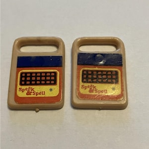 Peut inclure: Deux petits accessoires de jouet rectangulaires en plastique beige avec une poignée sur le dessus. Chaque pièce présente une bande bleue horizontale au-dessus d'un panneau jaune avec une grille noire et le texte rouge Speak & Spell.