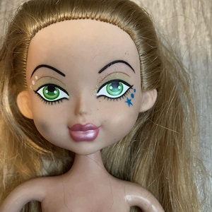 May include: A Bratz doll with green eyes, glitter eyeshadow, and a blue star tattoo. The doll has long blonde hair and pink lips. The doll's skin tone is light, and the doll is nude.