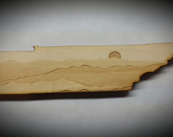 May include: A wooden cutout of the state of Tennessee with a sunset scene over mountains. The sun is a circle in the sky and the mountains are represented by lines.