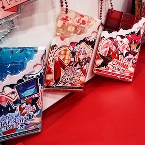 Könnte beinhalten: Drei dekorative, schokoladenriegelförmige Acryl-Anhänger. Die Anhänger zeigen farbenfrohe Illustrationen und Texte, darunter "Blue Raspberry Byte", "Candy Apple Creme" und "Red Cayenne Crunch". Jeder Anhänger ist an einer Perlenkette befestigt.