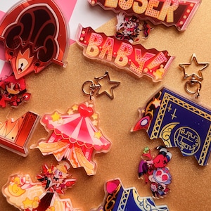 May include: A collection of acrylic keychains featuring various character illustrations and designs. The set includes pieces with the text LOSER BABY, a red circus tent, a blue book-shaped charm, and small character figures with star-shaped clasps.