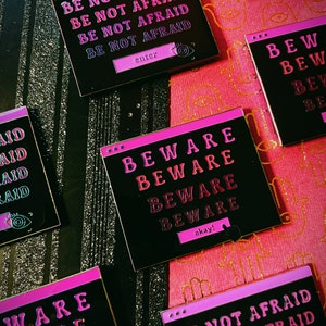以下が含まれることがあります： 「BE NOT AFRAID」と「BEWARE」のフレーズがレトロなコンピューターウィンドウのデザインで書かれた、黒とピンクの正方形のステッカー。ステッカーは光沢仕上げで、ピンクと黒の背景に配置されています。