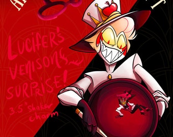 May include: Illustration featuring the text "AMERICAN VITCHCRAFT" and "Lucifer's Venison Surprise!" A character in a white outfit and a hat with a red apple. The background is red and black. A 8.89 cm shaker charm is also featured.