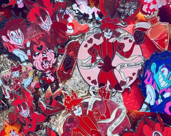 May include: A collection of enamel pins in various shades of red, pink, and gold. The pins feature cartoon characters and Valentine's Day themes, with some including text like "St. Valentine" and "May your heart have a frost."