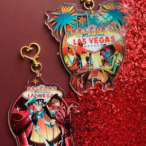 May include: Two acrylic keychains featuring cartoon characters and the text Welcome to Fabulous Las Vegas. One keychain has a gold star clasp, the other a gold heart clasp. The designs include vibrant colors, stage curtains, and palm trees.
